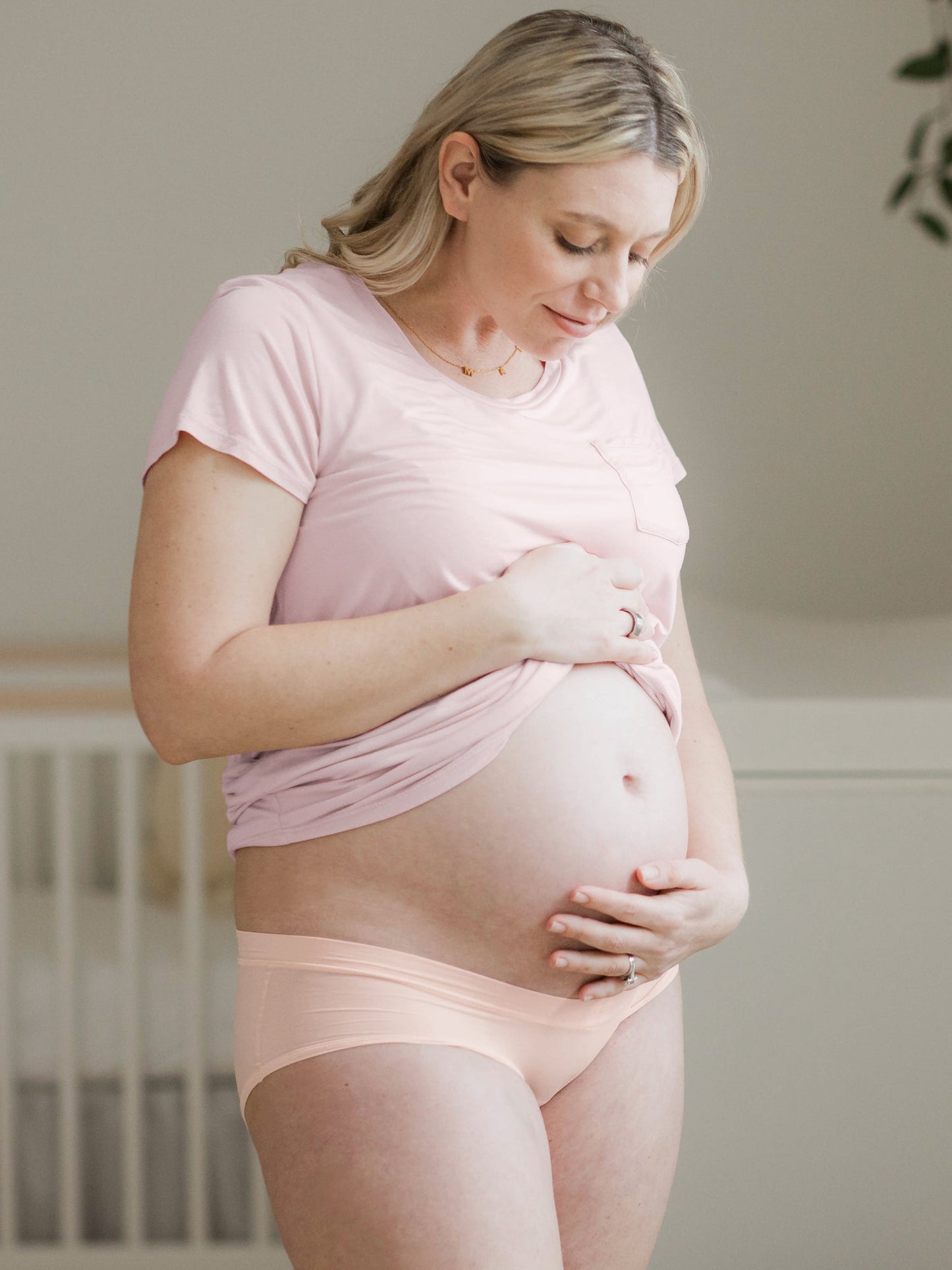 Model holding her baby bump while looking down and wearing the Grow with Me™ Maternity & Postpartum Hipster in Soft Pink 