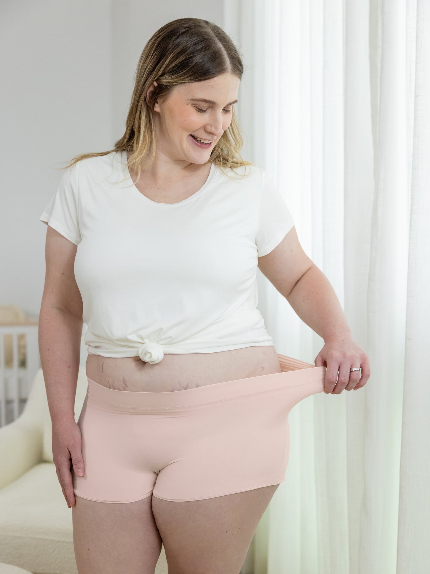 Model wearing the Everyday Nursing T-shirt in White tied up and paired with the Grow with Me™ Maternity & Postpartum Boyshort in Soft Pink. She is pulling the waistband of the underwear to show the stretch.