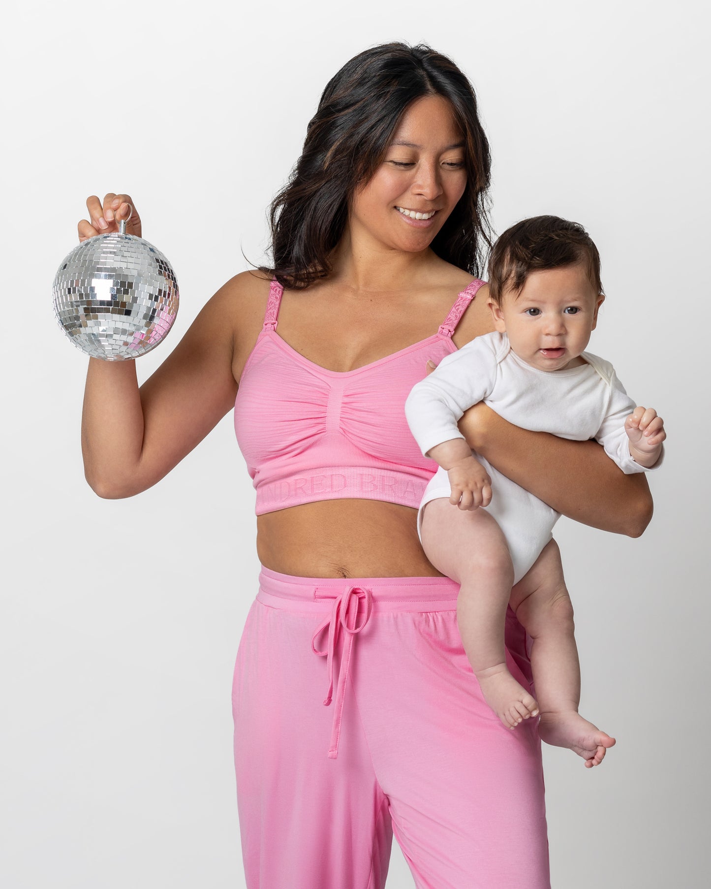 Model holding baby while wearing the Sublime® Hands-Free Pumping & Nursing Bra in Bubblegum with matching Everyday Lounge Joggers @model_info:Hillary is wearing a Small. 