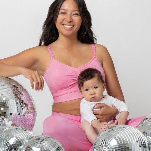 Model holding baby and sitting next to disco balls while wearing the Sublime® Hands-Free Pumping & Nursing Bra in Bubblegum with matching Everyday Lounge Joggers 