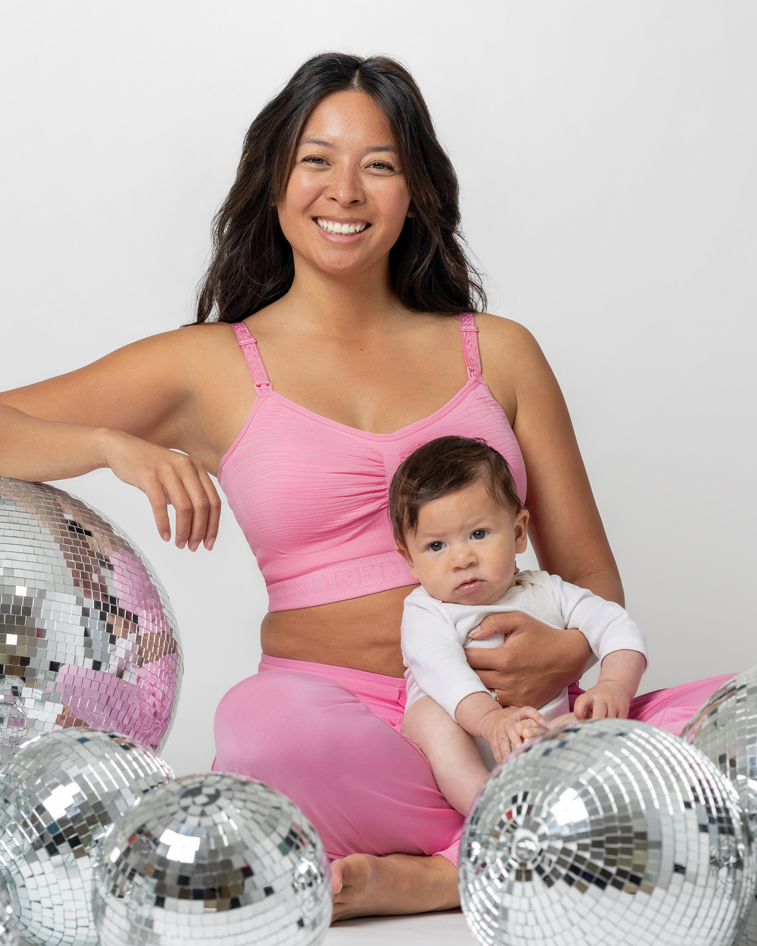 Model holding baby and sitting next to disco balls while wearing the Sublime® Hands-Free Pumping & Nursing Bra in Bubblegum with matching Everyday Lounge Joggers 