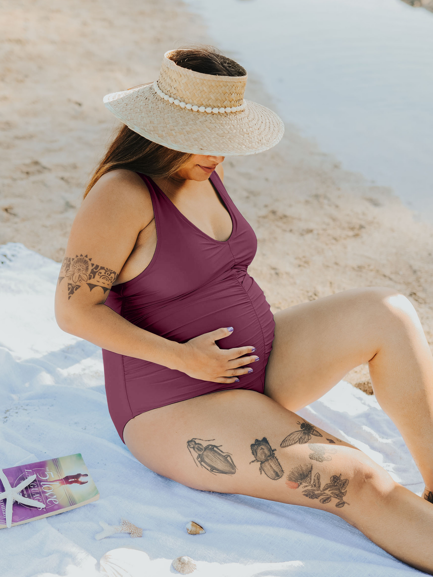 Pregnant model sitting on the beach wearing the Gathered Maternity One Piece Swimsuit in Maroon