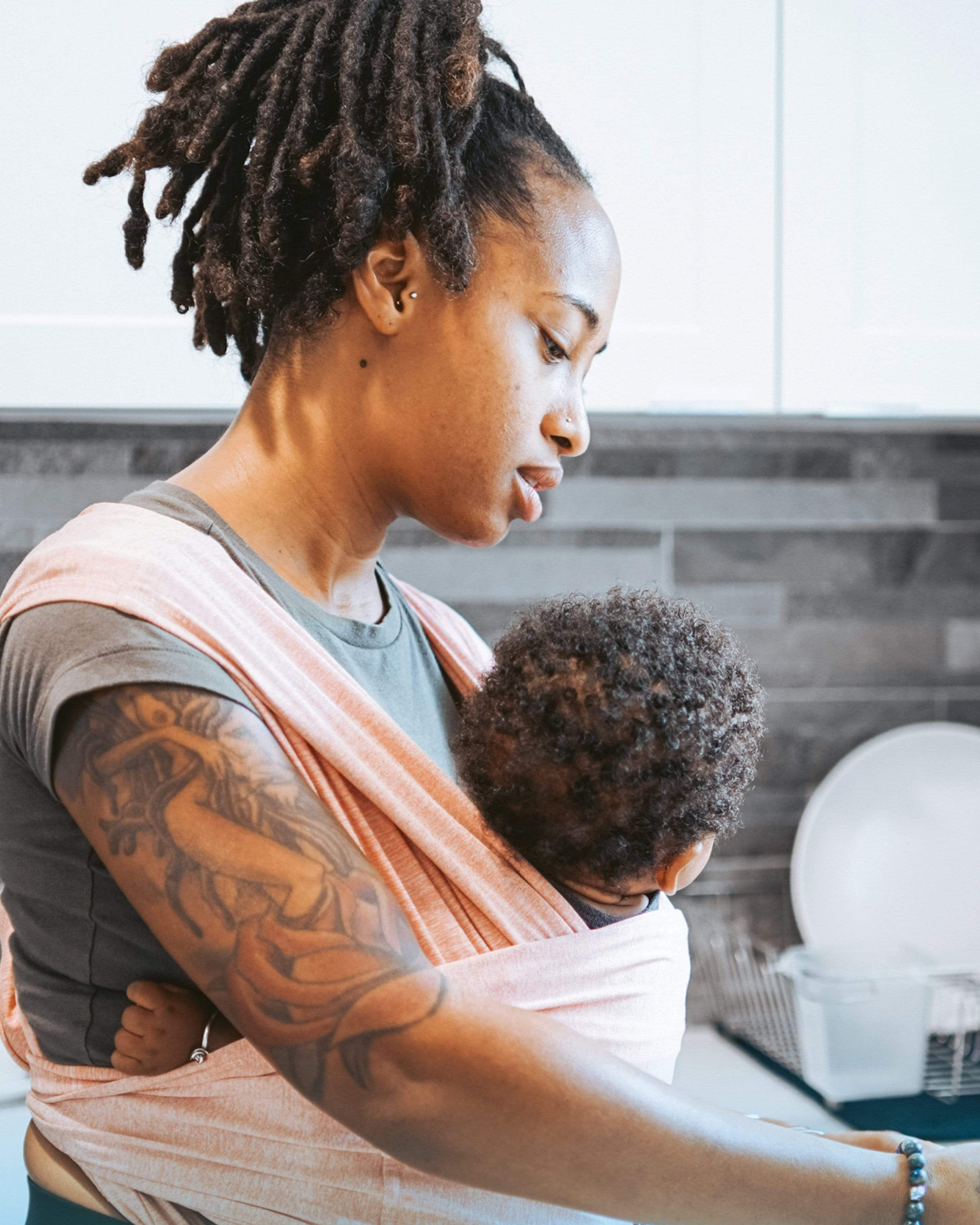 Model wearing baby in the Baby K'tan - Active Yoga Baby Carrier in Heather Coral while washing dishes