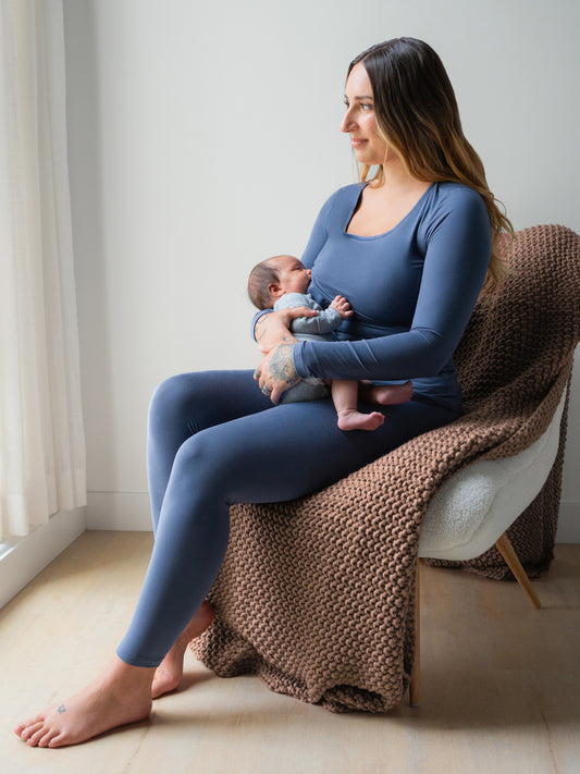 Model sitting in chair wearing the Jane Nursing Pajama Set in Slate Blue and nursing baby. @model_info:Anna is 5'6" and wearing a Small.