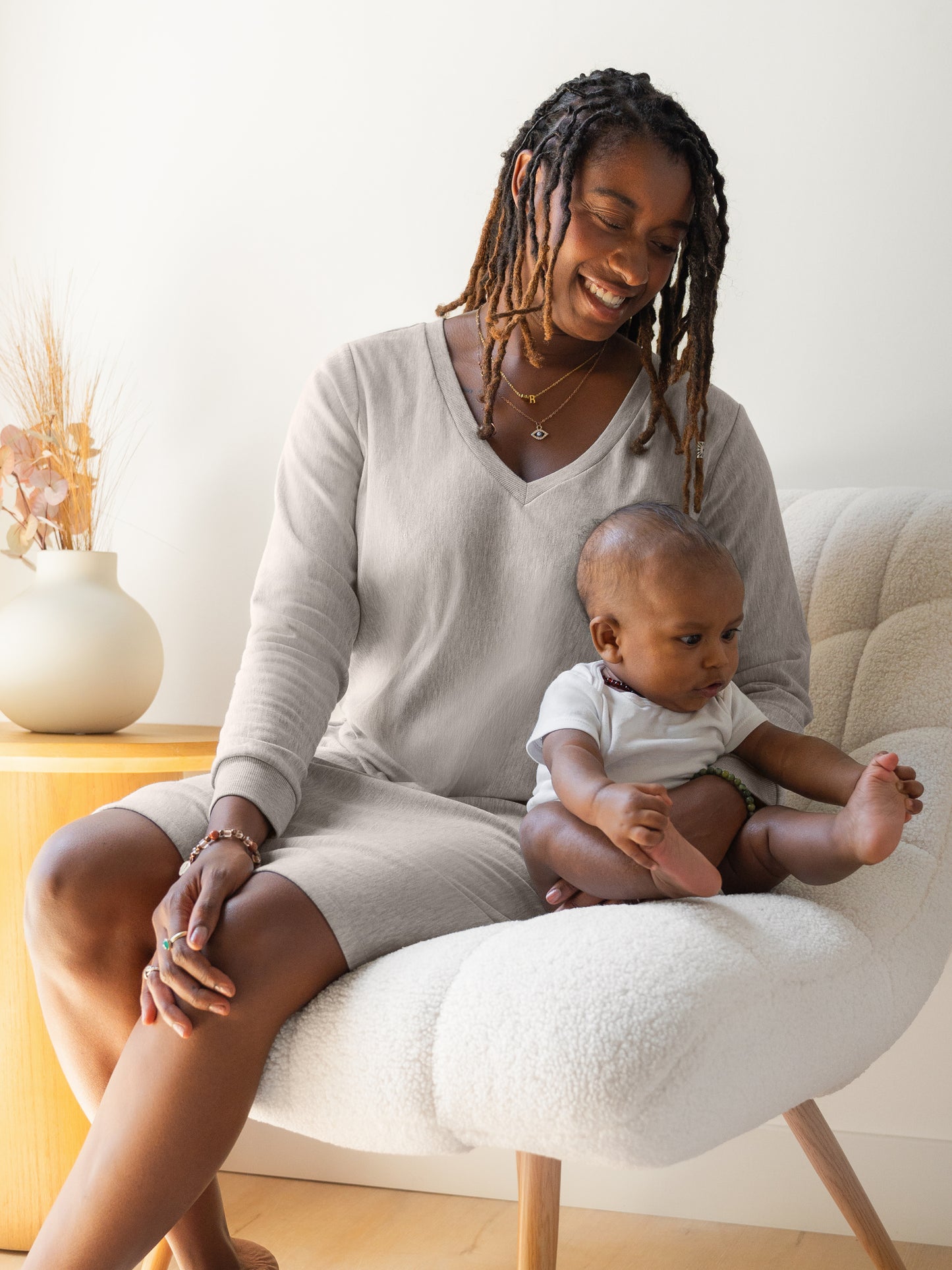 Model sitting on chair with baby, and wearing the Blake Oversized Maternity & Nursing Lounge Dress in Grey Heather