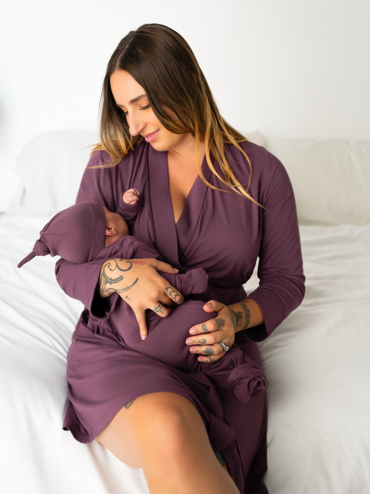 Model sitting on bed wearing the Clea Bamboo Robe in Burgundy Plum and cradling infant wearing the Baby Knotted Gown & Hat Set in matching Burgundy Plum. @model_info:Anna is 5'6" and wearing S/M.