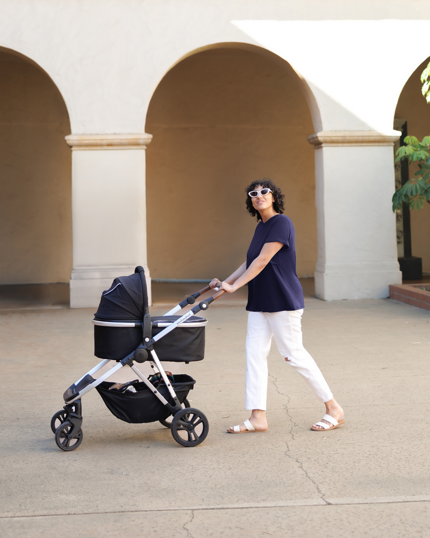 Model pushing stroller and wearing the Everyday Asymmetrical Nursing T-shirt in Navy, paired with cropped white jeans and sandals.