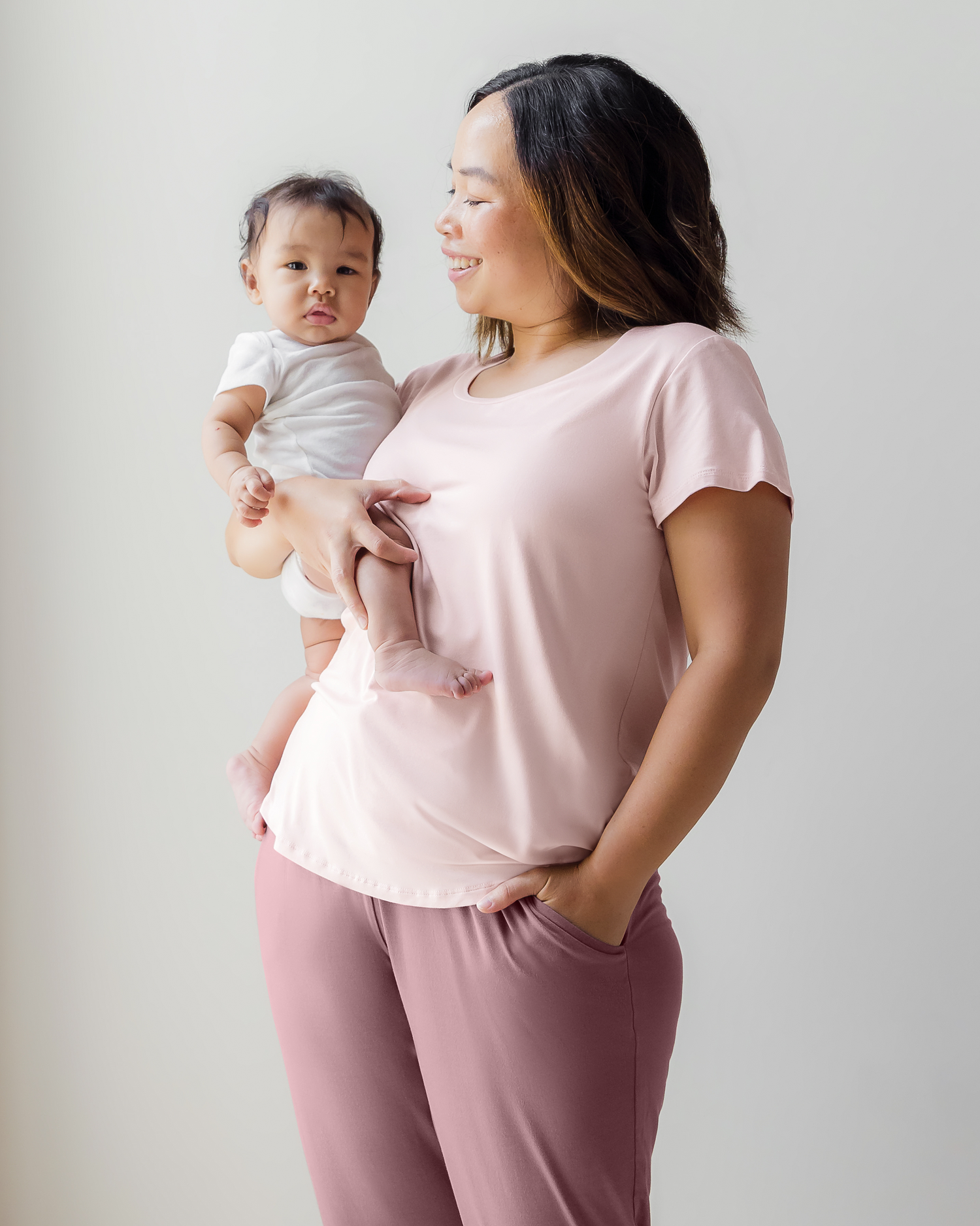 Model wearing the Everyday Nursing T-shirt in Pink paired with Everyday Lounge Jogger in Twilight, holding baby in one arm