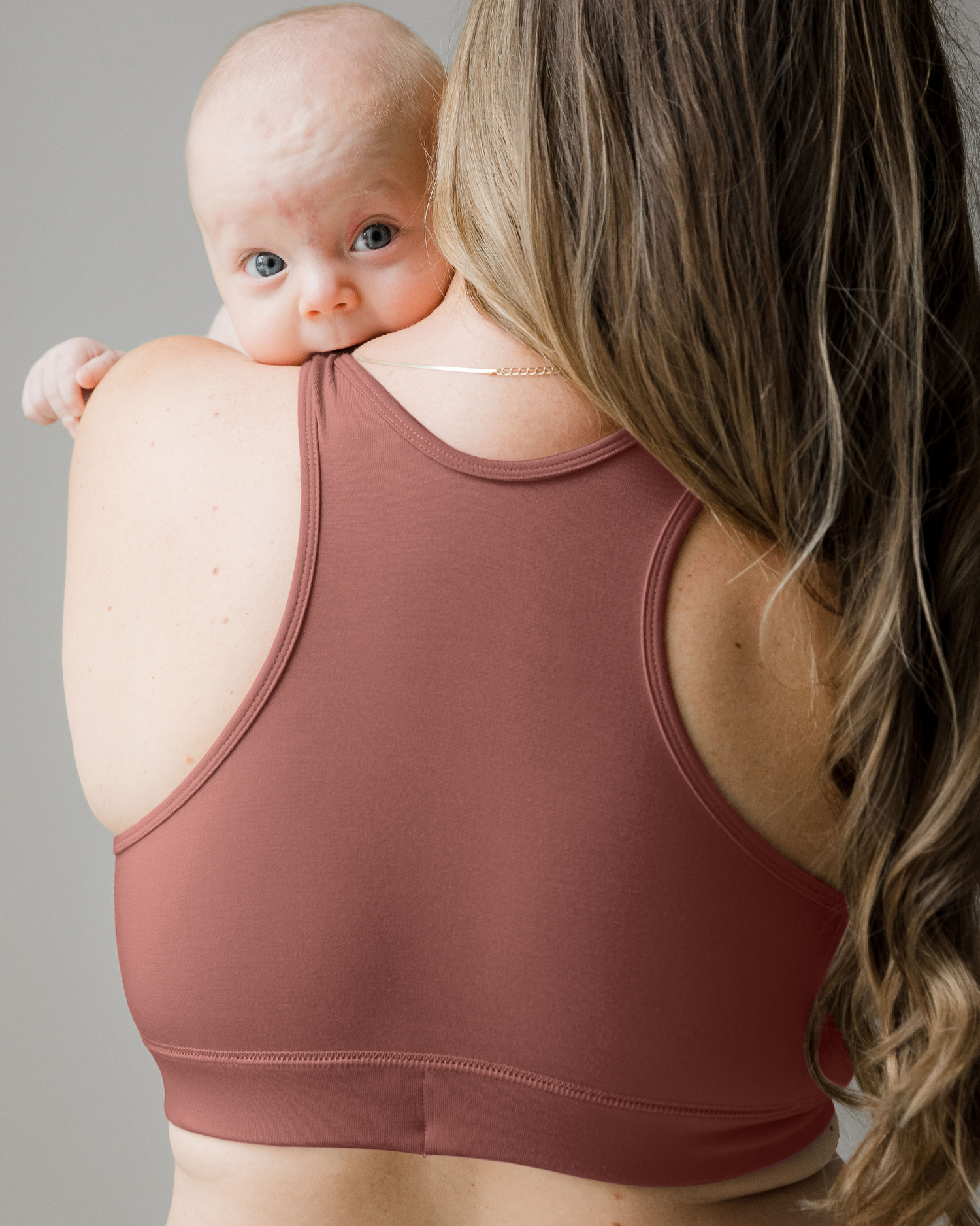 Back view of model wearing the French Terry Racerback Nursing & Sleep Bra in Redwood holding her baby who is peeking over her shoulder