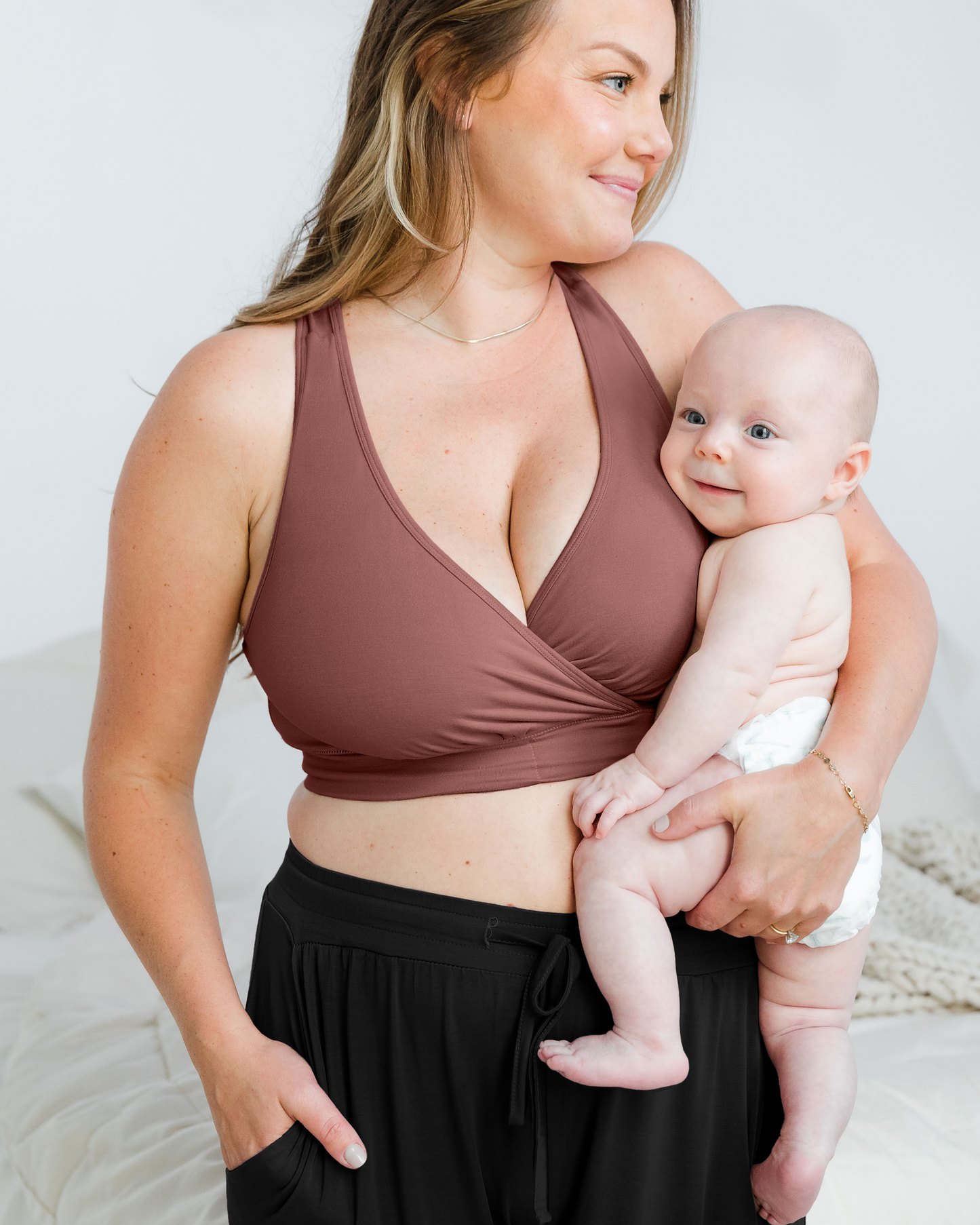 Model wearing the French Terry Racerback Nursing & Sleep Bra in Redwood holding her baby against her chest. @model_info:Ali is wearing a Large Busty.