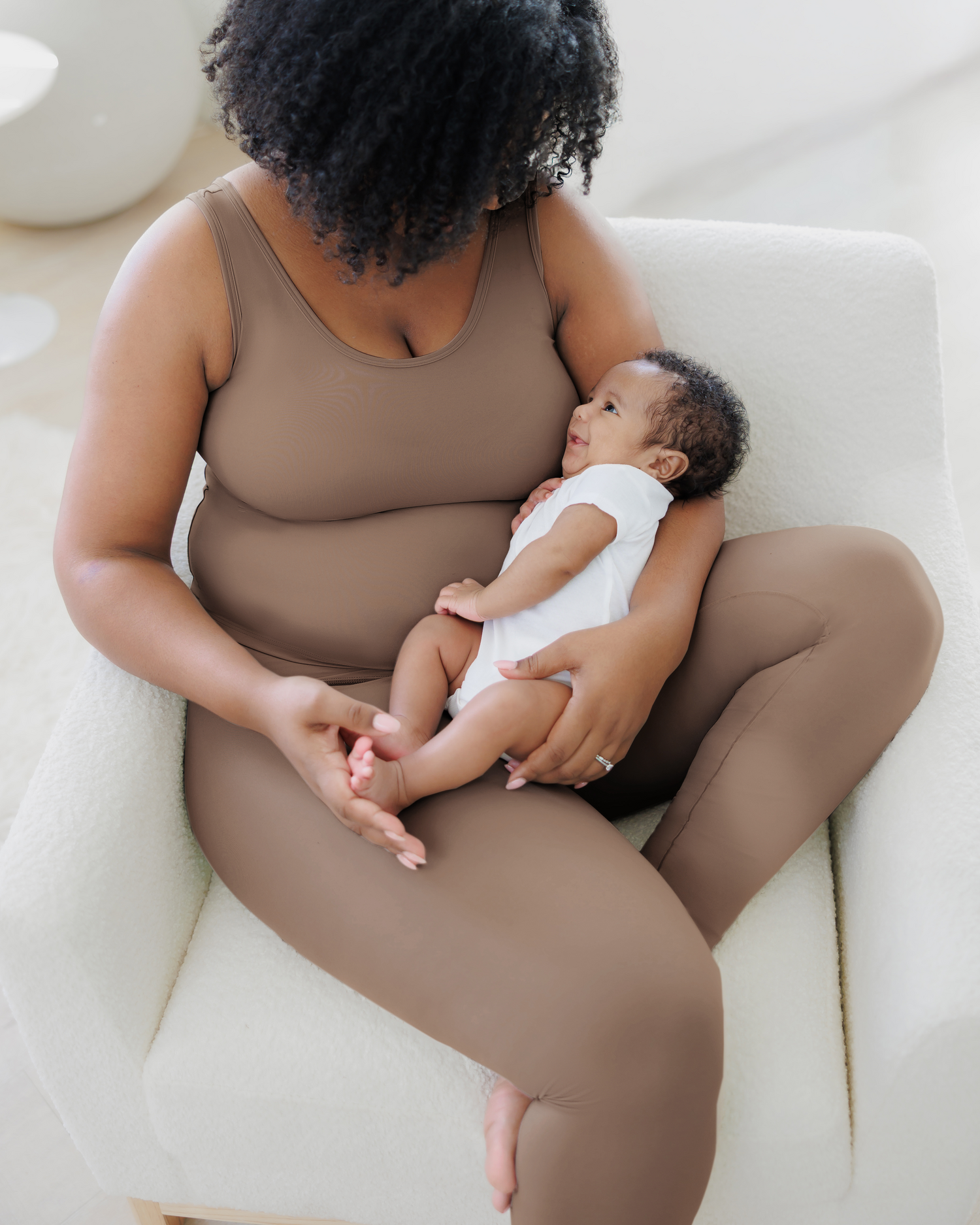 Model sitting in chair cradling baby and wearing the Grow with Me™ Maternity & Postpartum Legging in Taupe paired with matching Grow with Me™ Maternity & Nursing Tank