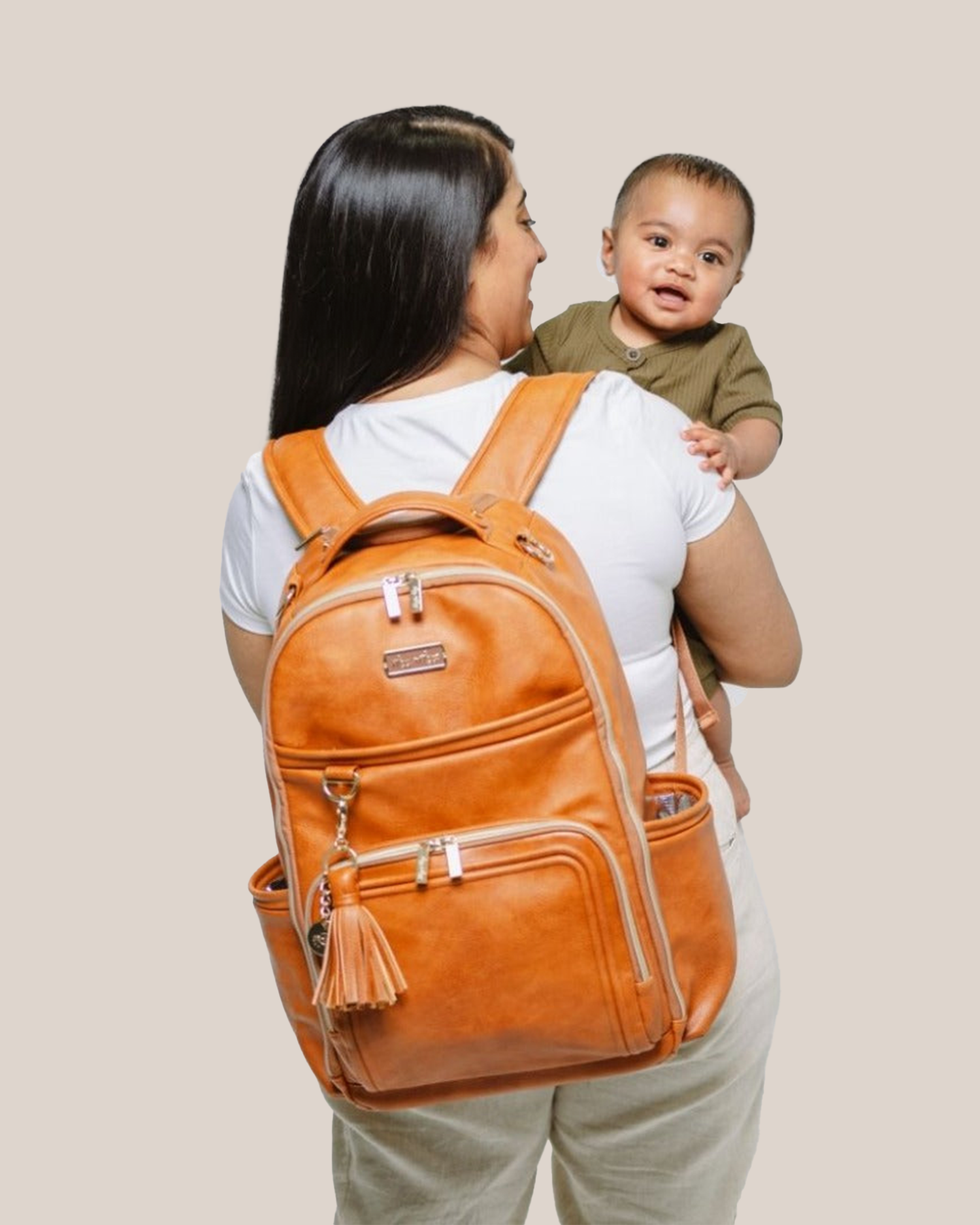 Model wearing the Itzy Ritzy - Boss Plus™ Diaper Bag Backpack in Cognac, holding baby