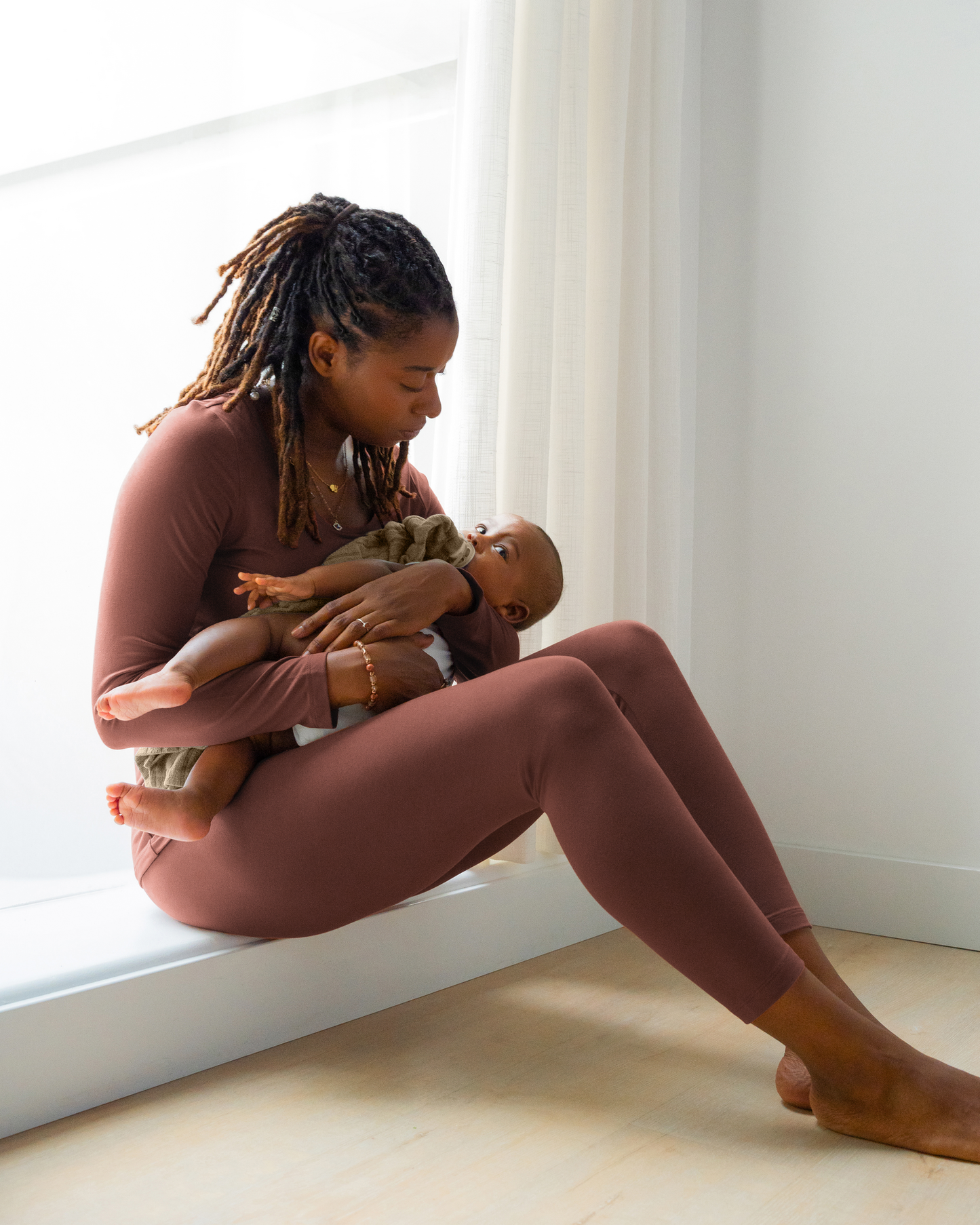 Model sitting wearing the Jane Nursing Pajama Set in Redwood, and cradling baby.