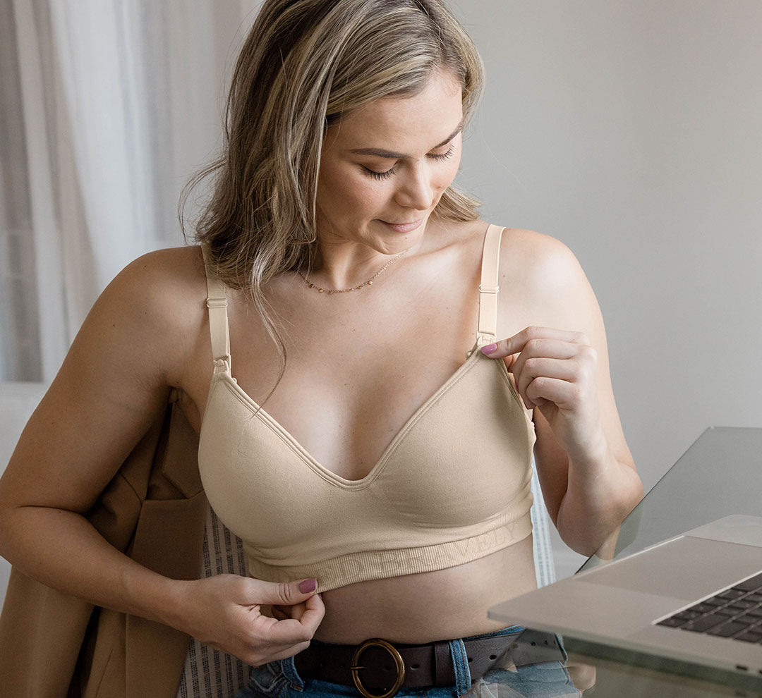 Model sitting at desk wearing the Signature Sublime® Contour Hands-Free Pumping & Nursing Bra in Beige, showing clip-down access