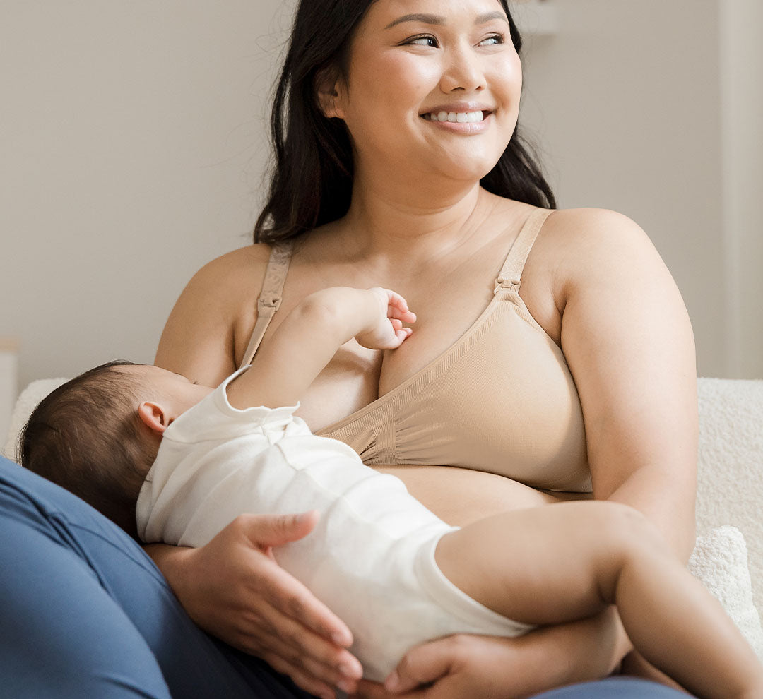 Model sitting in chair wearing the Simply Sublime® Nursing Bra in Beige and nursing baby