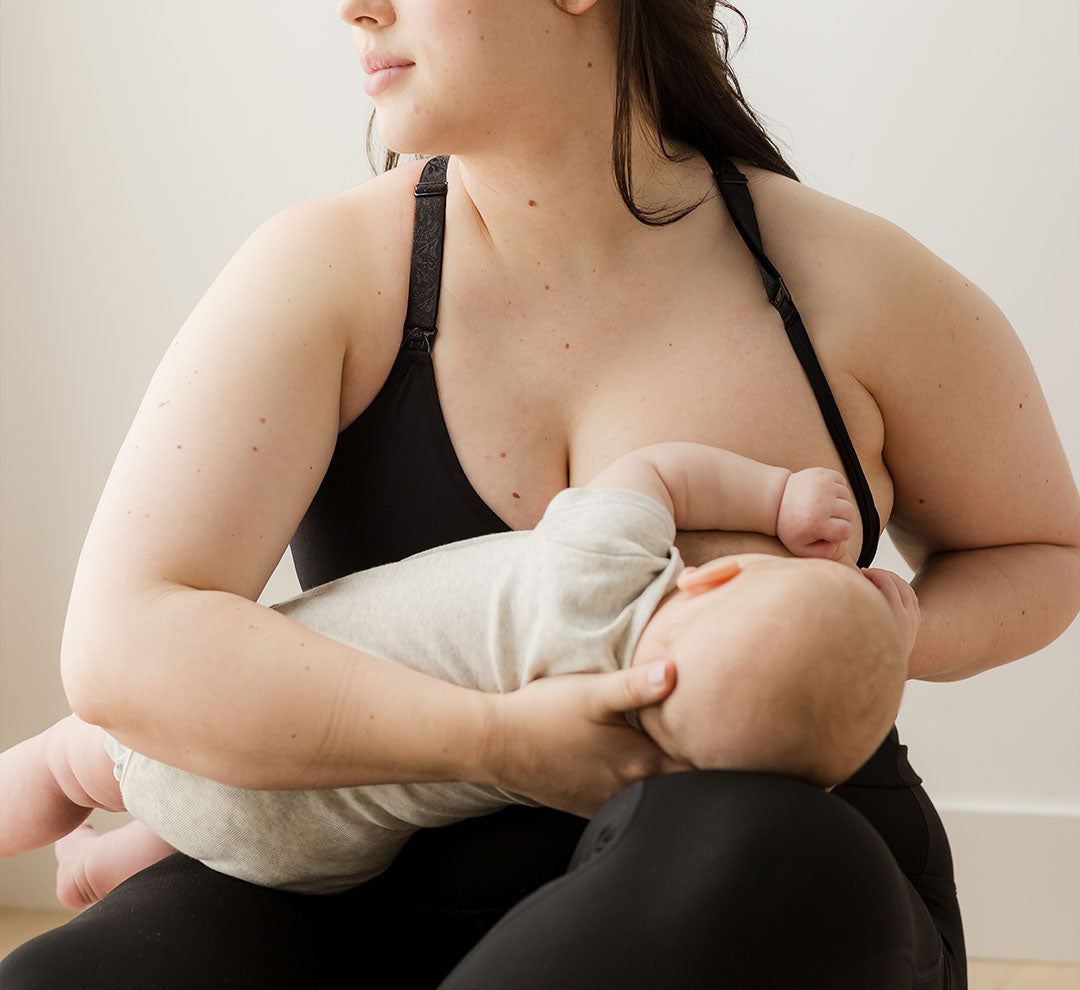 Model wearing the Sublime® Nursing Sports Bra in black, sitting and nursing baby