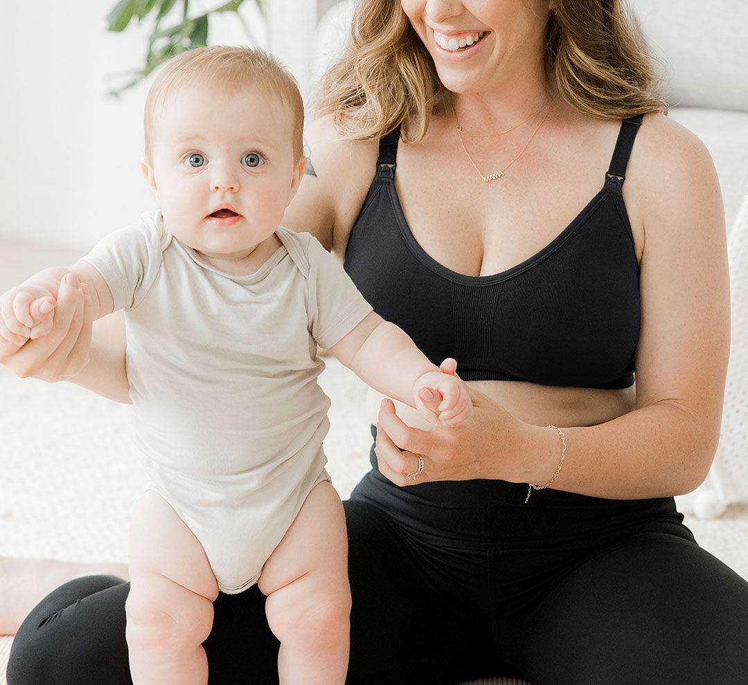 Model wearing the Sublime® Hands-Free Pumping & Nursing Sports Bra in black, holding hands of baby who is standing in front of her