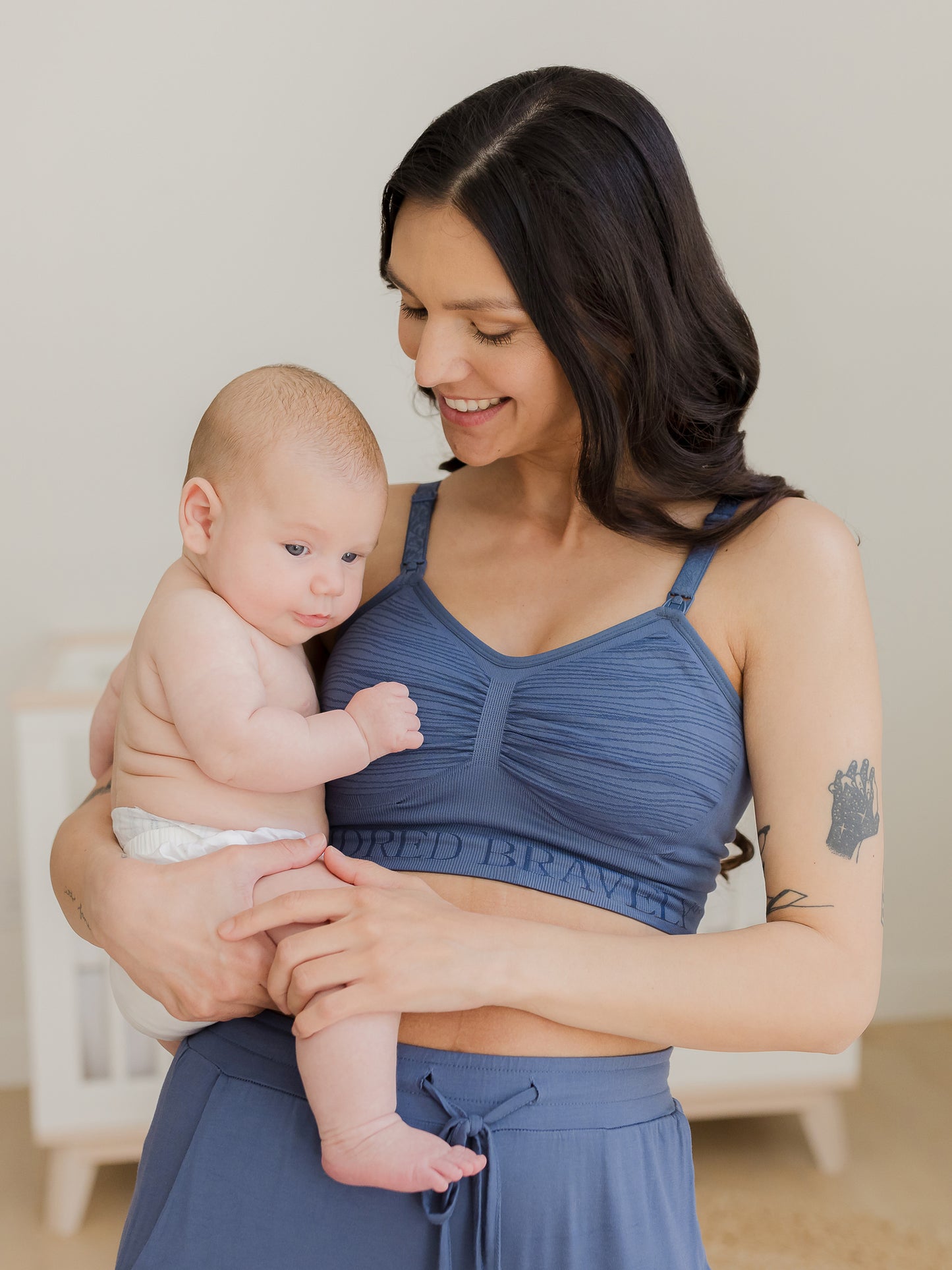 Model standing in nursery wearing the Sublime® Hands-Free Pumping & Nursing Bra in Slate Blue and holding baby.