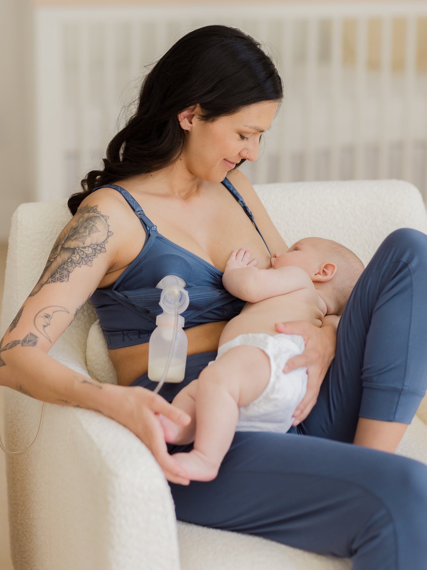 Model sitting in chair wearing the Sublime® Hands-Free Pumping & Nursing Bra in Slate Blue, nursing baby and pumping at the same time. @model_info:Megan is wearing a Small.