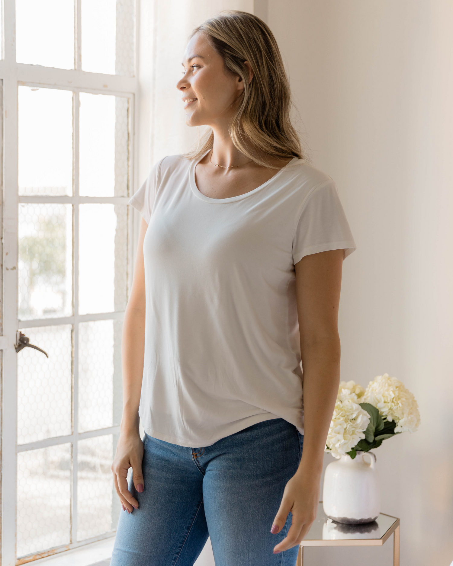 Model standing by a window wearing the Everyday Maternity & Nursing T-shirt in White @model_info:Taylor is wearing a Medium.