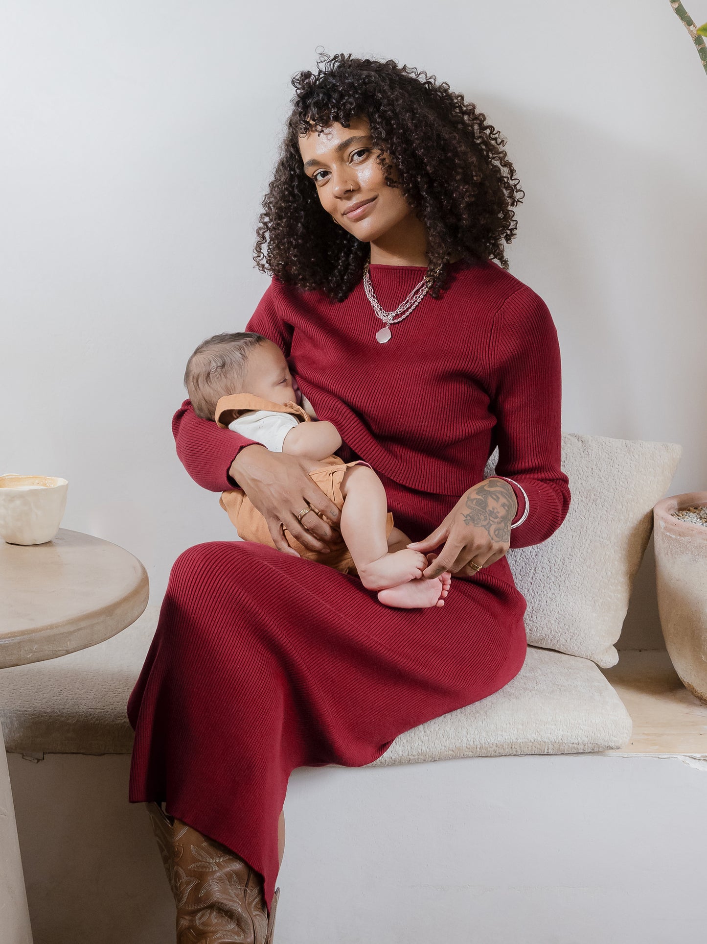 Model sitting in cafe wearing the 2-in-1 Maternity & Nursing Midi Dress in Garnet nursing baby @model_info:Rudi is 5'8" and wearing a Small.