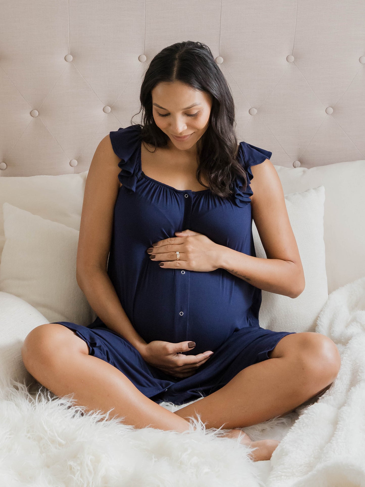 Pregnant model sitting cross legged on bed wearing the Ruffle Strap Labor & Delivery Gown in Navy. @model_info:Julana is wearing an XS/S.