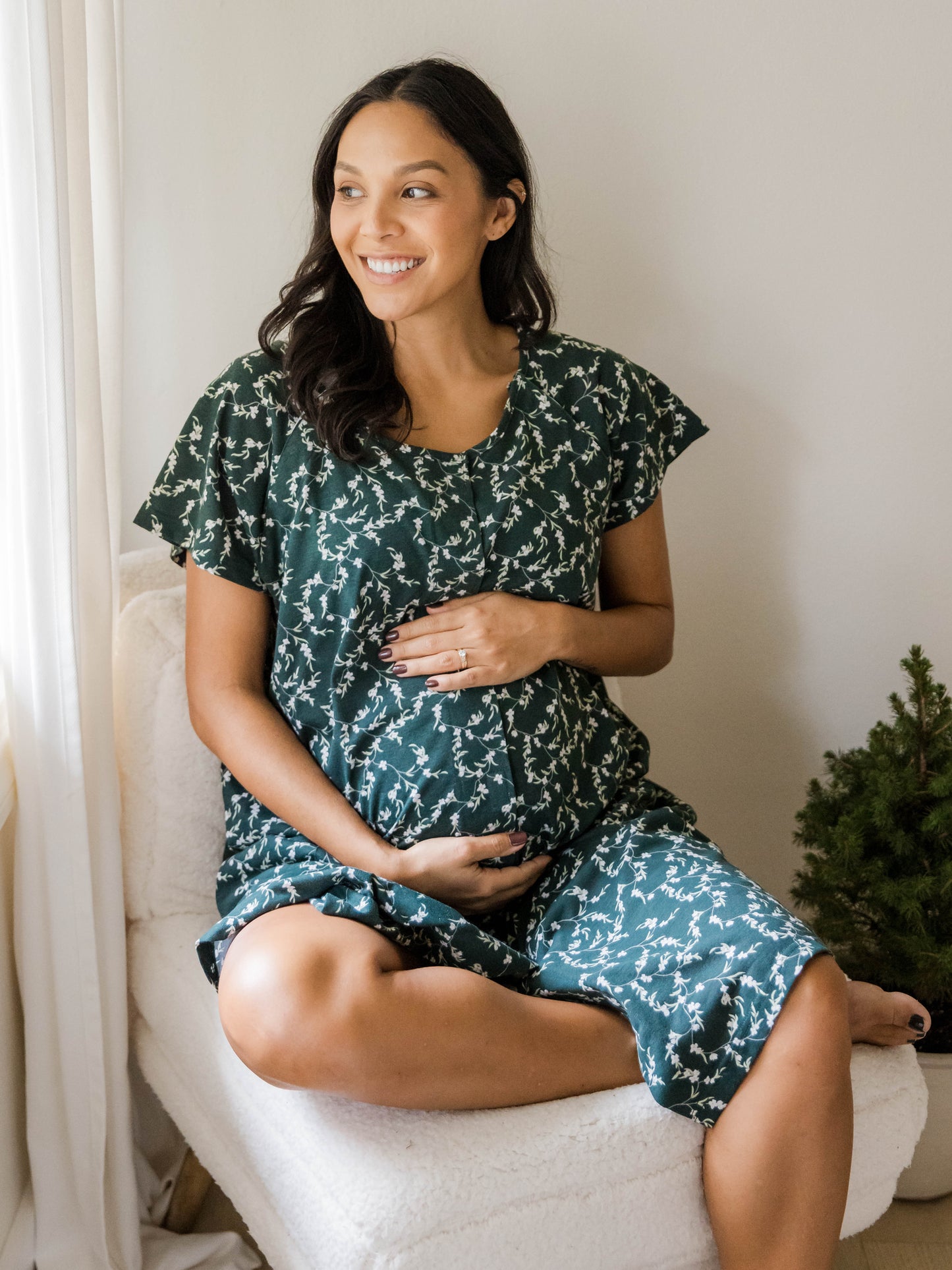 Pregnant model sitting in chair wearing the Universal Labor & Delivery Gown in Evergreen Blossom. @model_info:Julana is wearing a S/M/L.