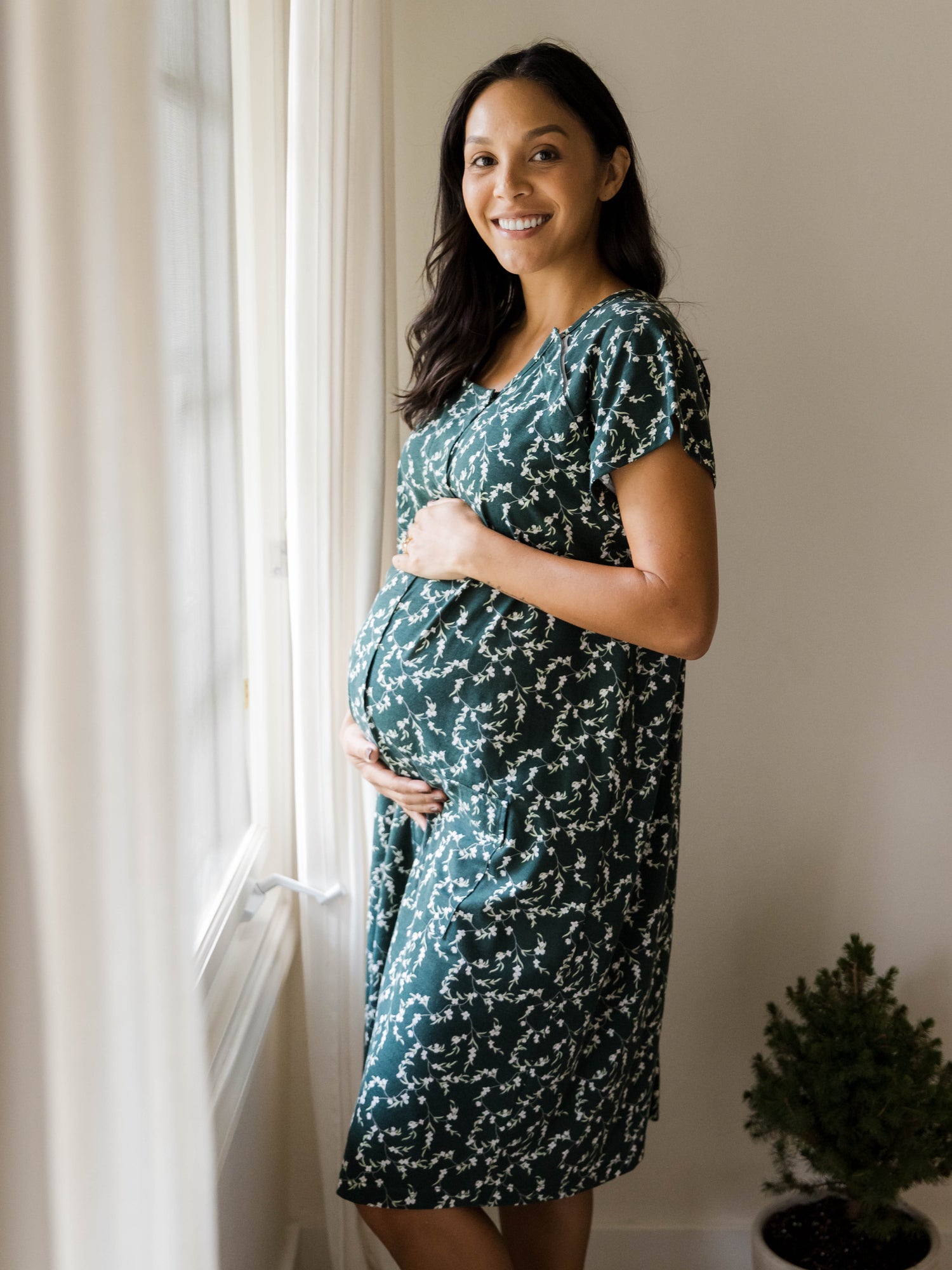 Pregnant model standing near window wearing the Universal Labor & Delivery Gown in Evergreen Blossom.