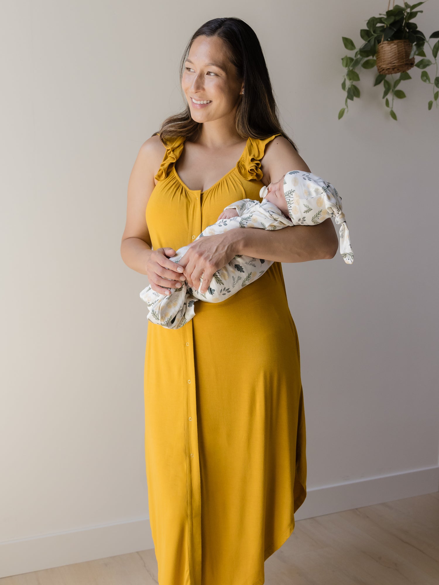 Model wearing the Ruffle Strap Labor & Delivery Gown in Honey holding baby wearing the Baby Knotted Gown & Hat Set in Honey Dandelion.