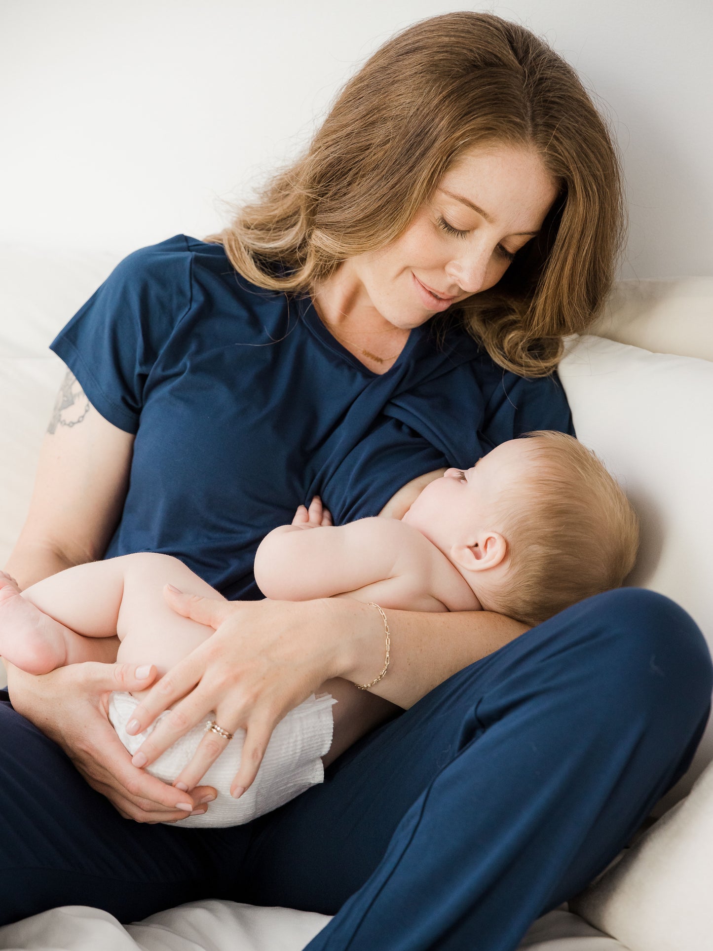 Model sitting on bed nursing baby and wearing a Short Sleeve Tulip Hem Maternity Pajama set - Top & Bottom in Navy Blue.