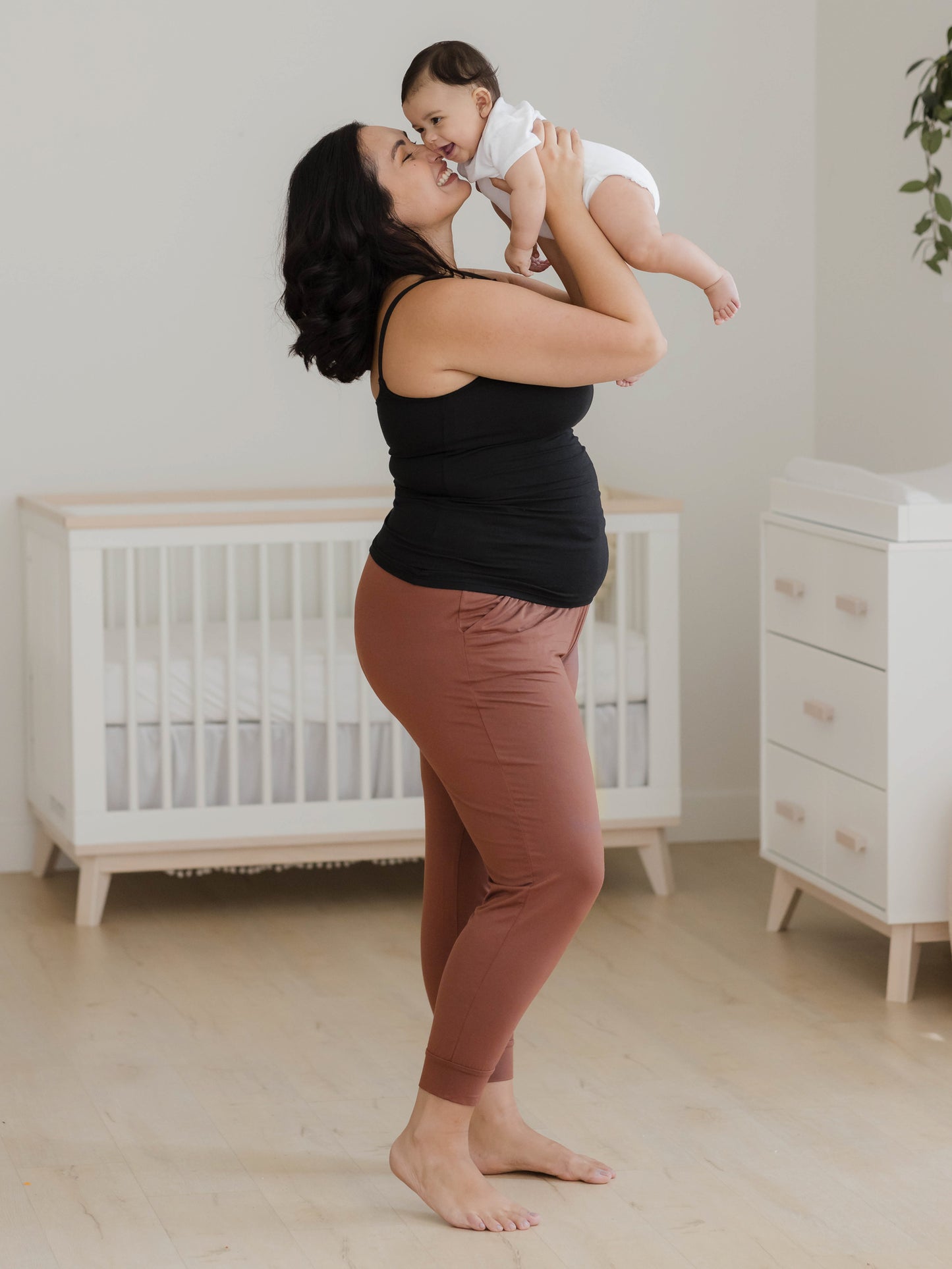 Full body side view of model holding baby above her face wearing the Picot Trim Nursing Camisole in Black, paired with the Everyday Lounger Jogger in Redwood.