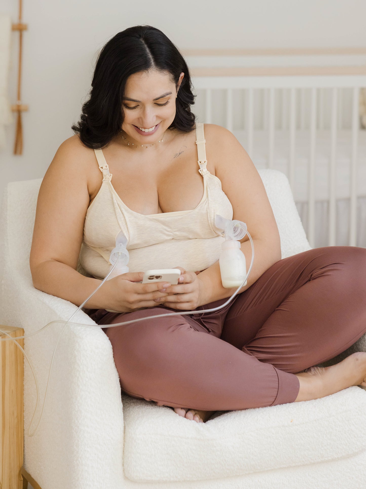  Model sitting in chair pumping and holding phone and wearing the Sublime® Bamboo Hands-Free Pumping & Nursing Camisole in Oatmeal Heather