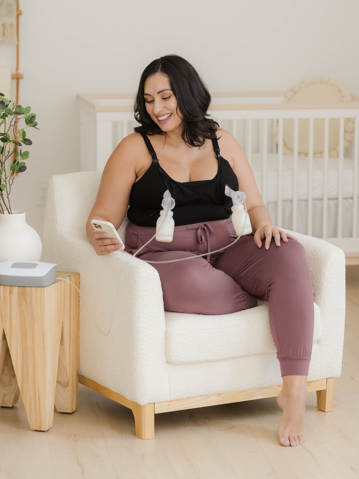 Model sitting in chair pumping and holding phone and wearing the Sublime® Bamboo Hands-Free Pumping & Nursing Camisole in Black