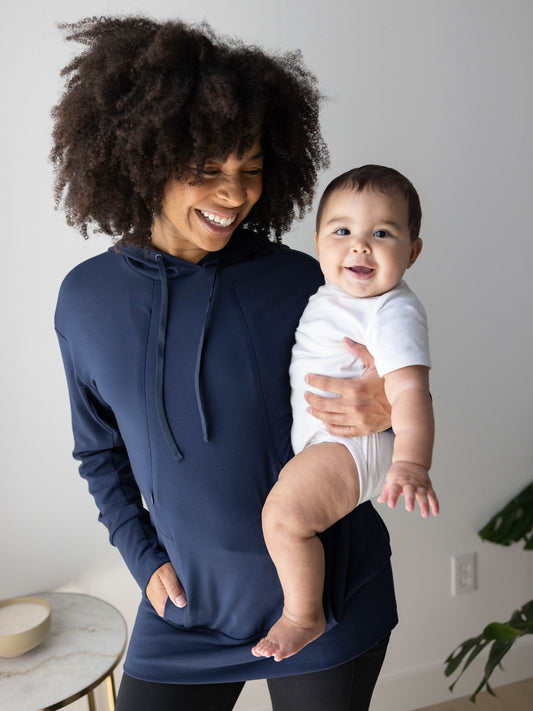 Front view of model wearing the Bamboo Maternity & Nursing Hoodie in Navy holding baby. @model_info:Tess is wearing a Small.