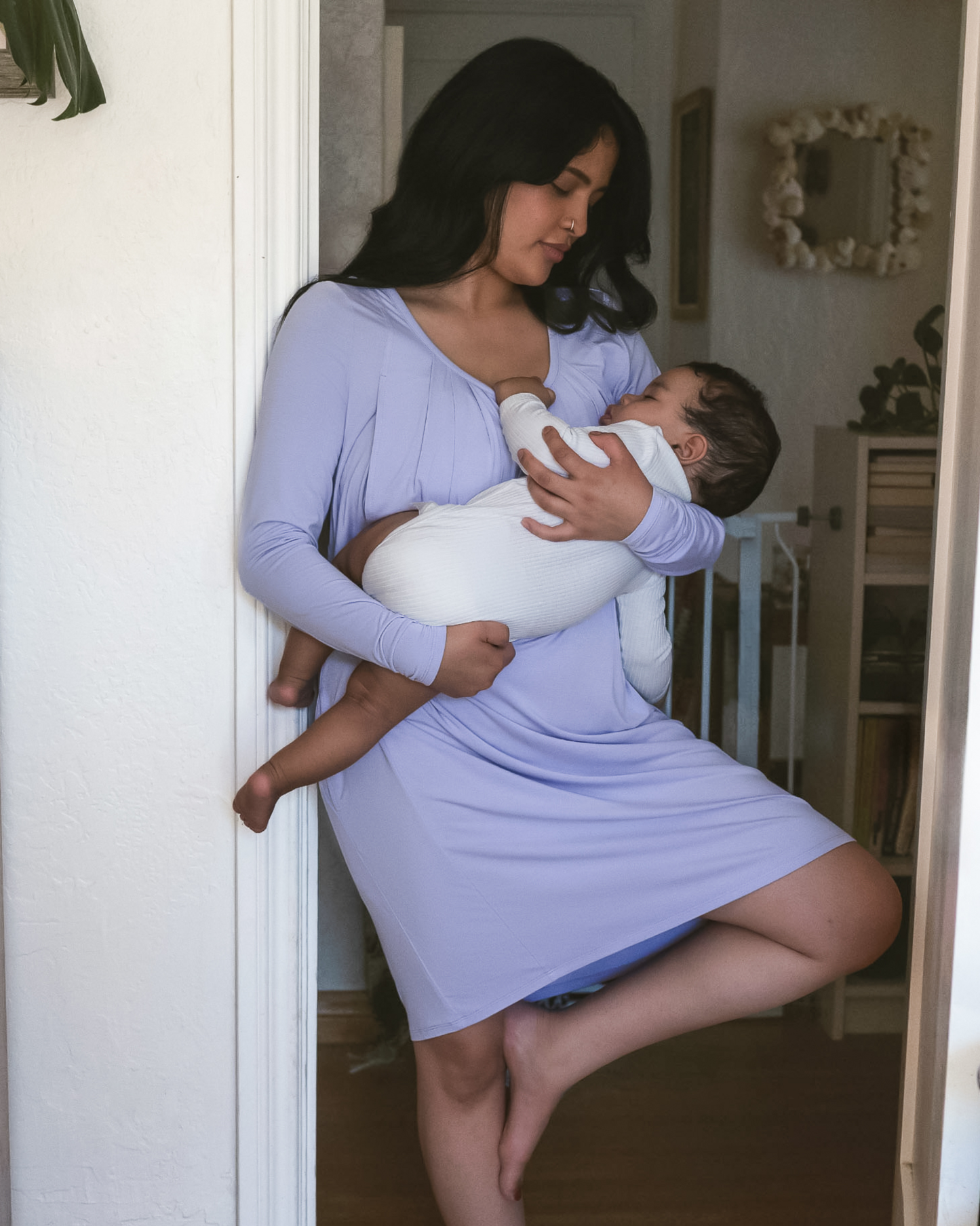 Model leaning against doorway wearing the Quinn Bamboo Long Sleeve Maternity & Nursing Nightgown in Periwinkle, cradling sleeping baby @model_info:Betsabe is wearing a Medium.