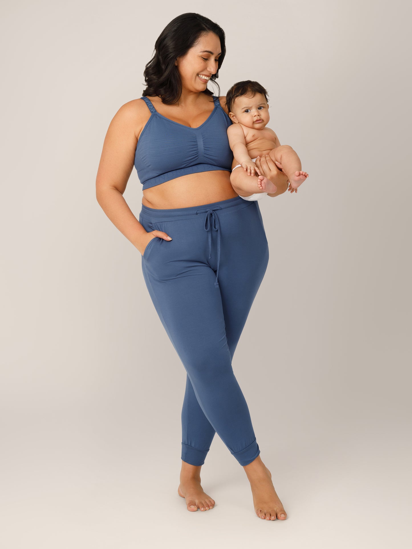 Front view of model wearing the Sublime® Hands-Free Pumping & Nursing Bra in Slate Blue in a Busty fit paired with matching Everyday Lounge Jogger, and holder baby in one arm.