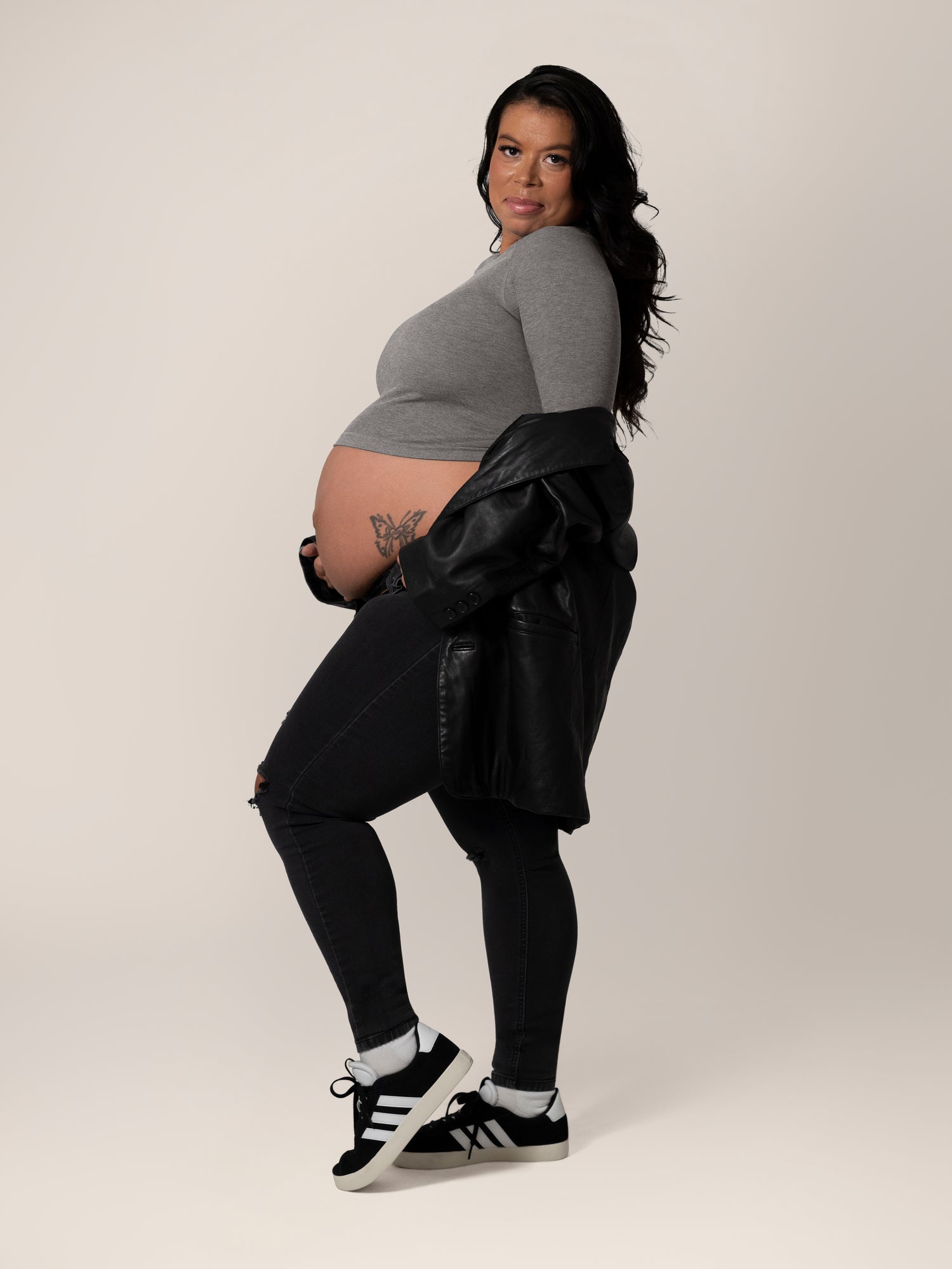 Full body side view of model wearing the Sublime® Bamboo Maternity & Nursing Cropped Long Sleeve Top in Charcoal, paired with black jeans and jacket hanging down on arms.