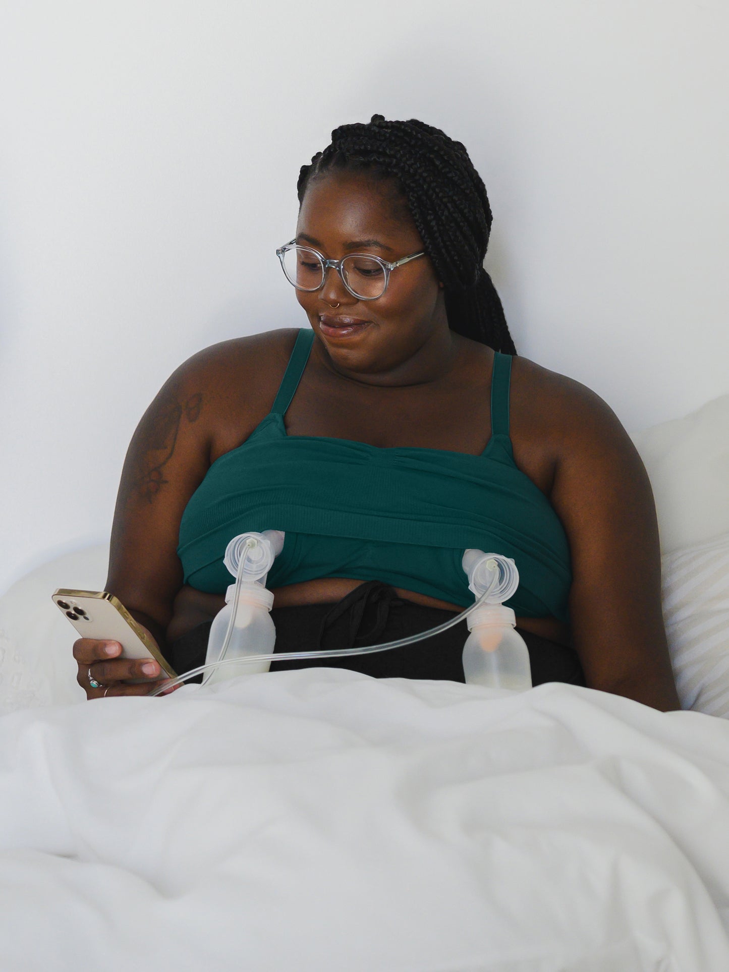 Model wearing the Sublime® Bamboo Hands-Free Pumping Lounge & Sleep Bra in Evergreen, pumping in bed and looking at phone. @model_info:Leondra is wearing a 1X Busty.