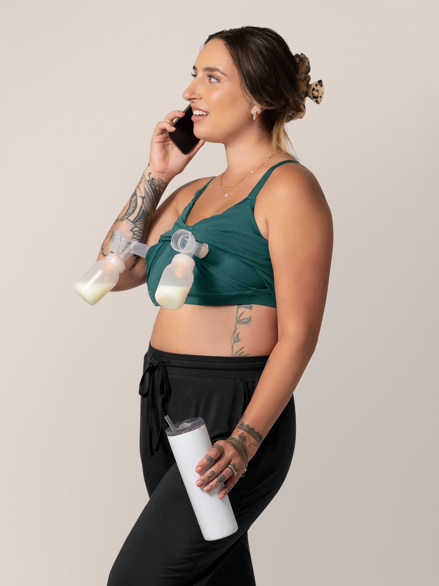 Front view of model wearing the Sublime® Bamboo Hands-Free Pumping Lounge & Sleep Bra in Evergreen, with two pump flanges inserted, while holding phone in one hand and water bottle in the other