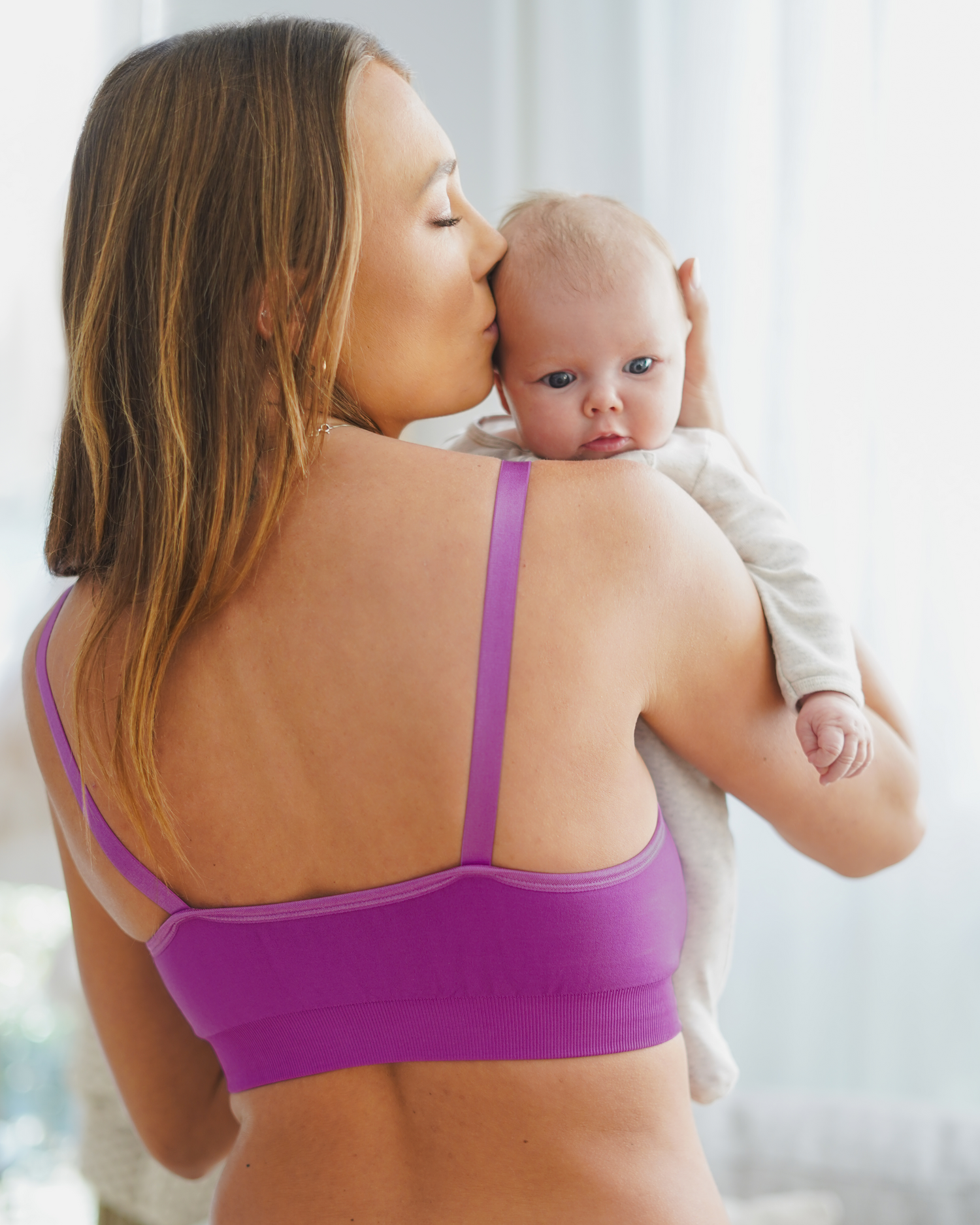 Back view of model wearing the Tatum Seamless Maternity & Nursing Bralette in Orchid, holding and kissing baby