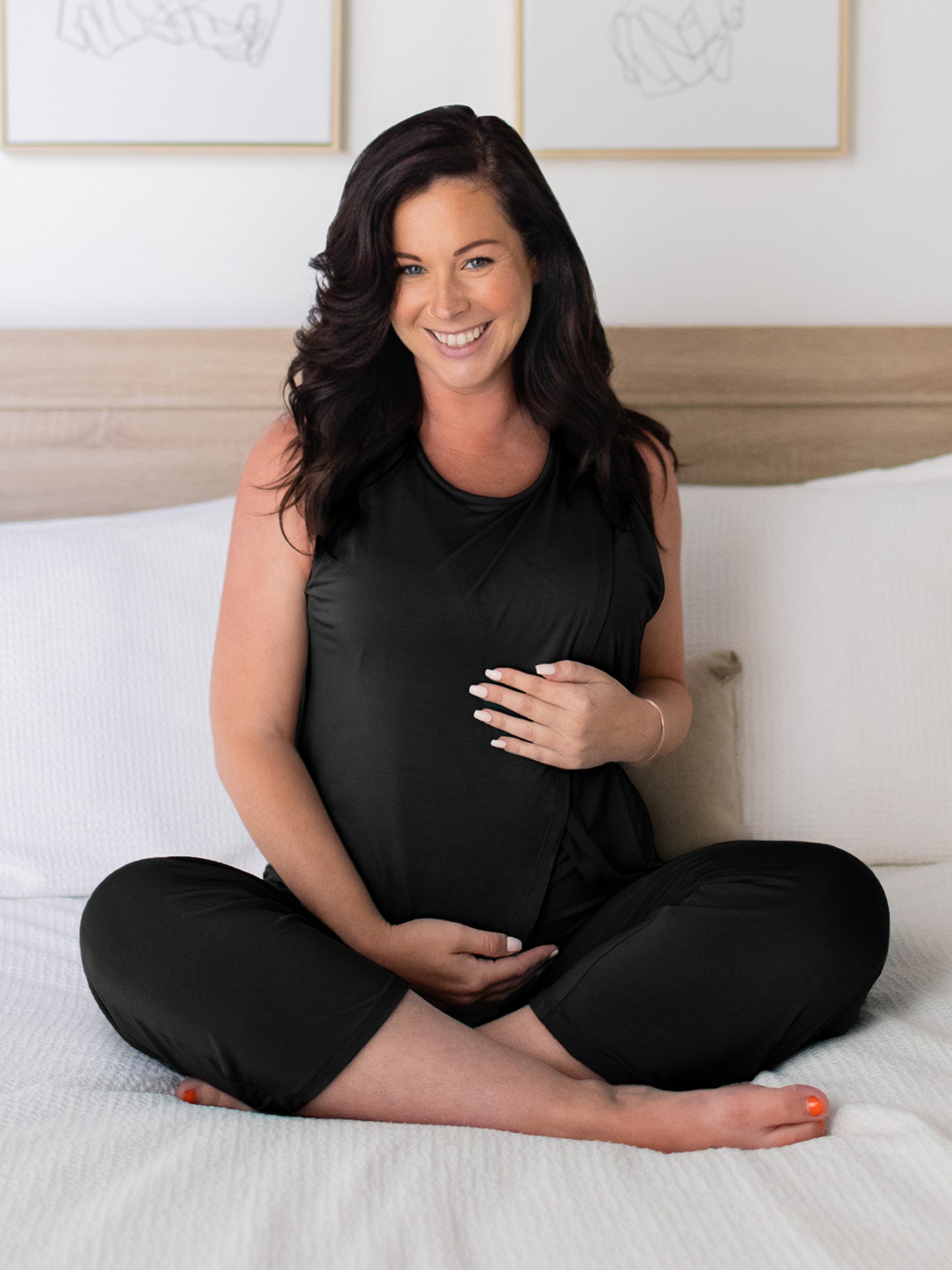 Front view of pregnant model sitting cross-legged on bed wearing the Tulip Hem Nursing Tank & Capri Pajama Set in Black.